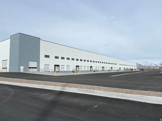 Industrial facility featuring a white and gray exterior, numerous dock-height loading doors, multiple ground-level roll-up doors, an expansive paved lot, and a mountain backdrop