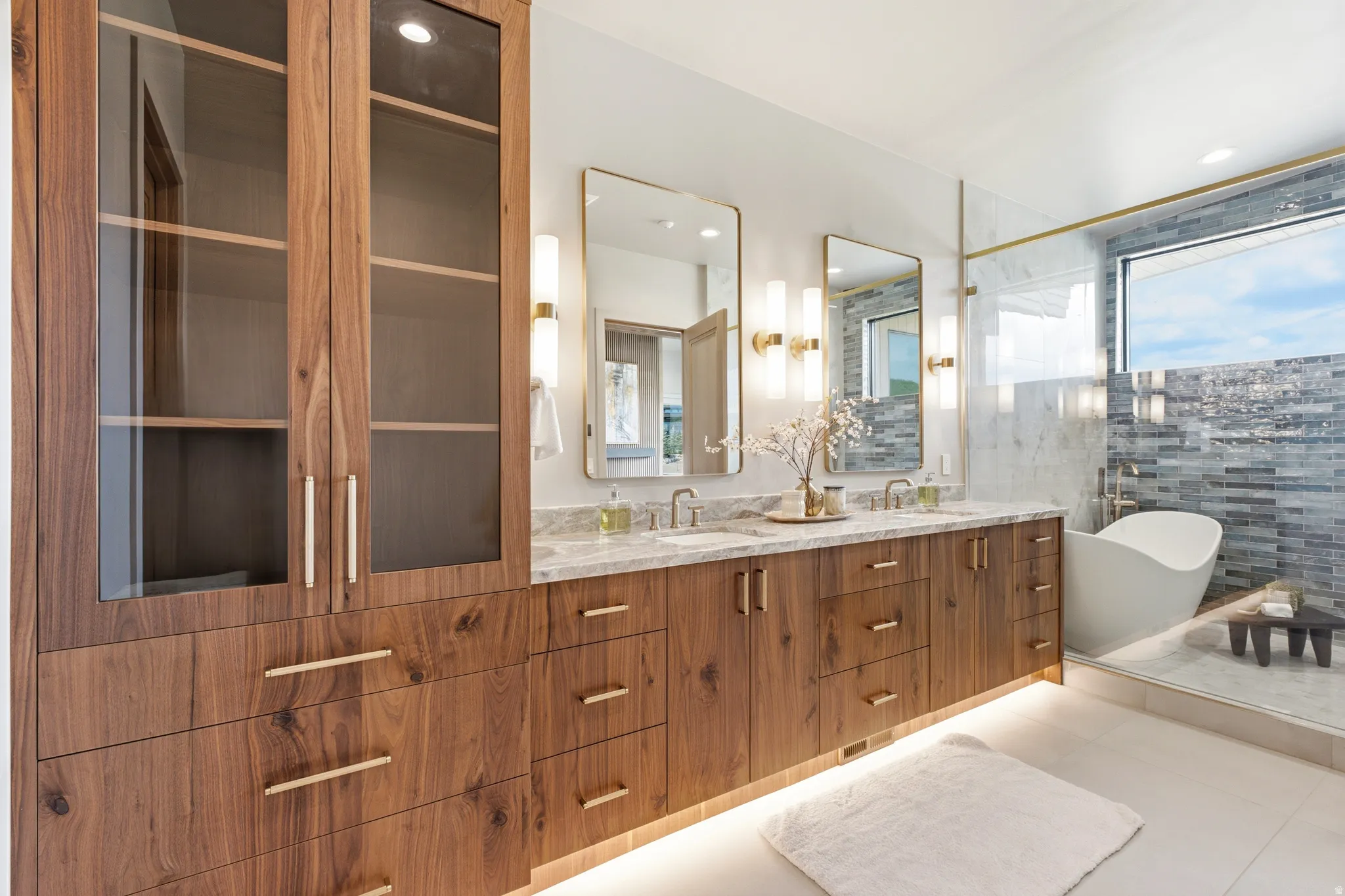 Custom wood-finish cabinetry with glass-front uppers, double vanity with stone countertops, dual rectangular mirrors, and a freestanding soaking tub