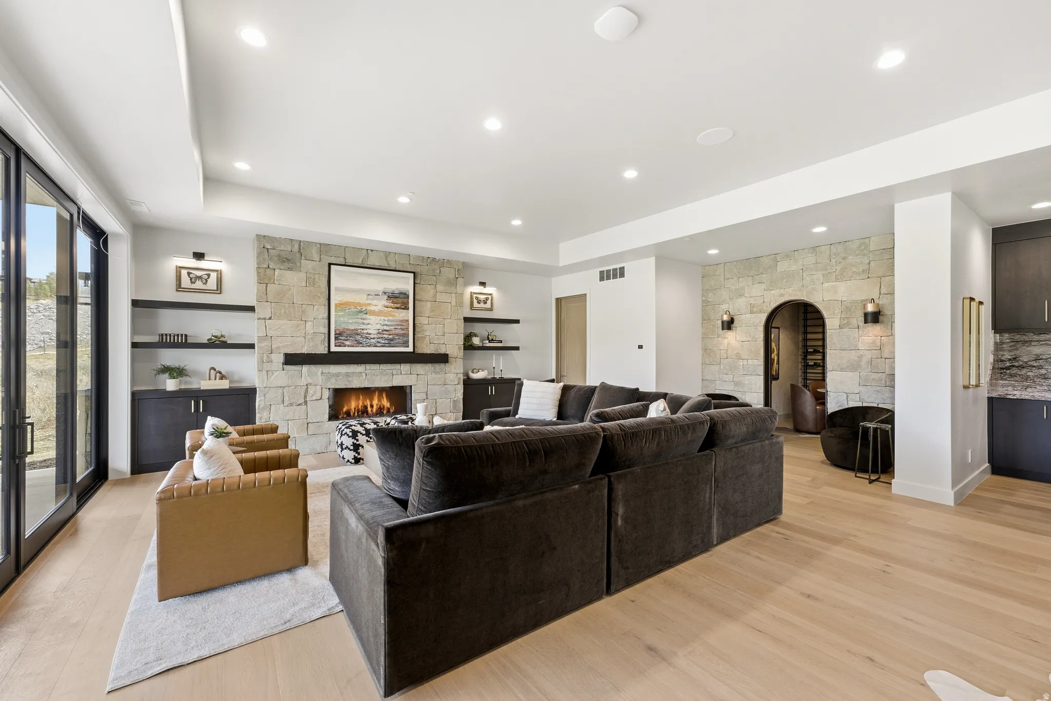 Expansive living area featuring light wood-finish flooring, a stone-clad fireplace with a dark wood mantel, built-in shelving, and sliding glass doors