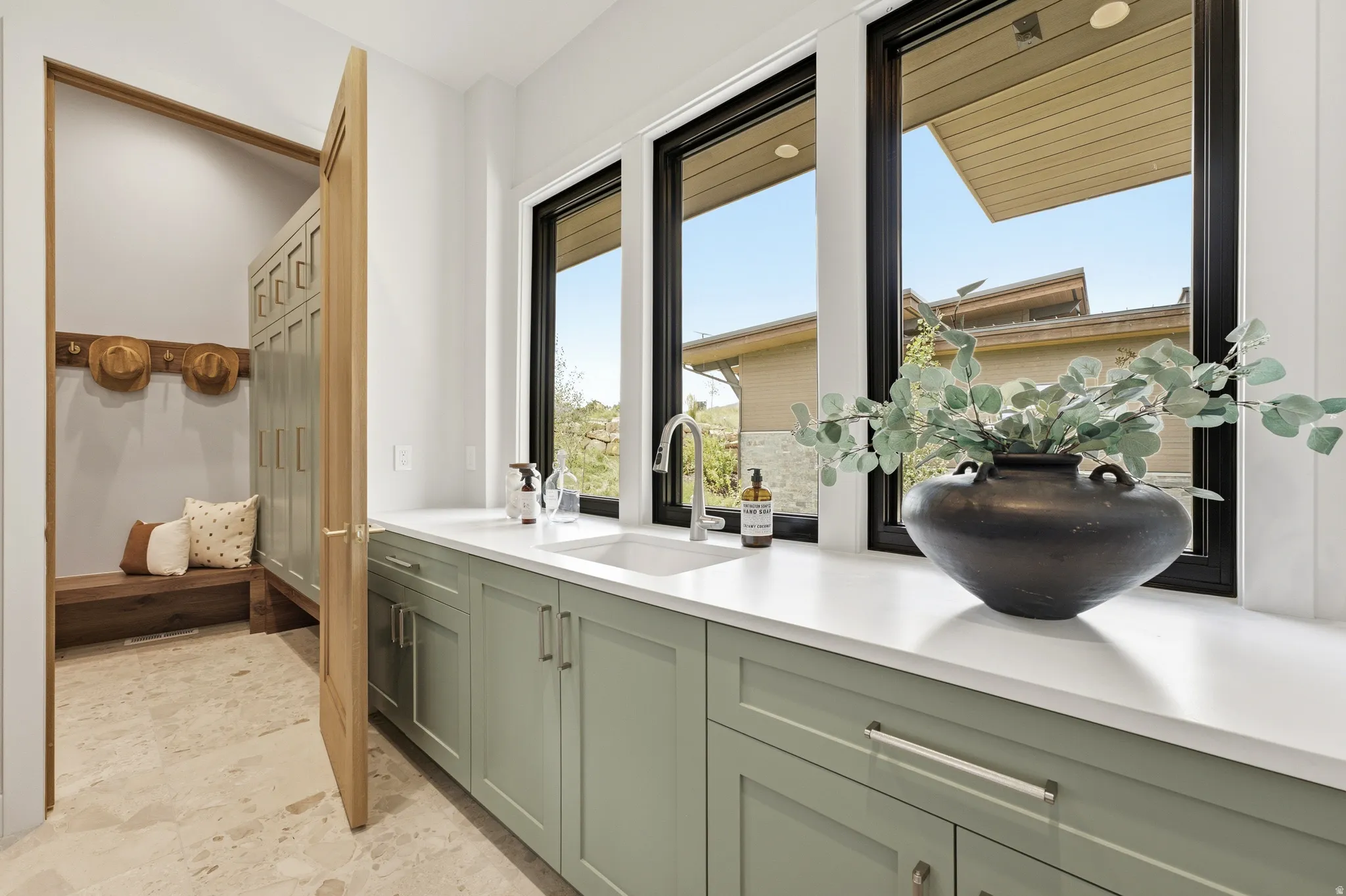 Built-in mudroom bench with wood-finish hooks, custom green cabinetry, white countertop with an integrated sink, and black-framed windows