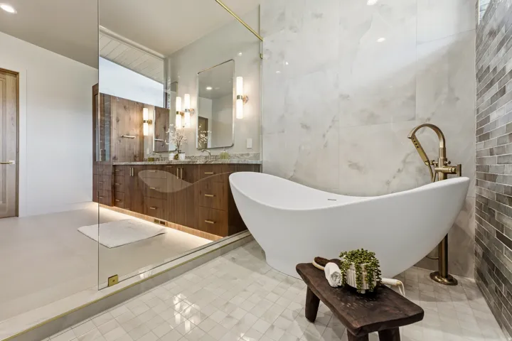 Freestanding soaking tub with floor-mounted faucet, glass-enclosed shower, wood-finish double vanity with undermount sinks, and wall-mounted sconce lighting