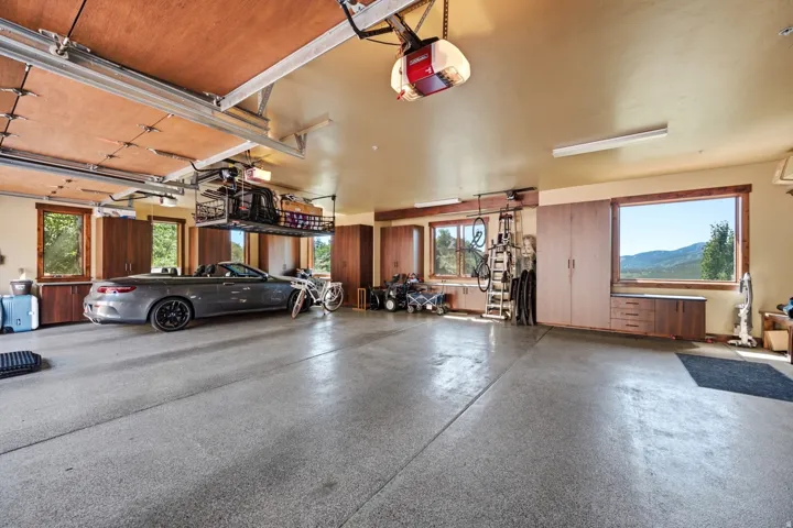 Spacious garage featuring a durable epoxy floor, extensive built-in cabinetry, multiple windows with natural light, overhead storage racks, and fluorescent lighting