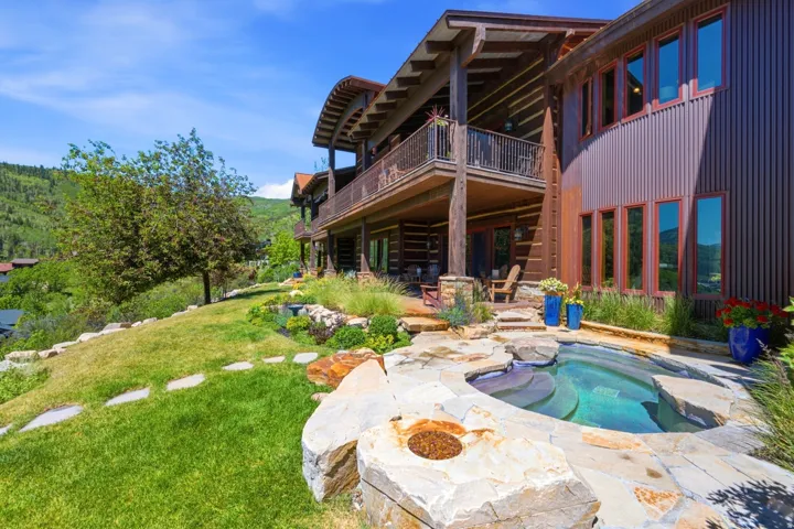 Multi-level exterior featuring a stone-surrounded spa with integrated fire pit, expansive lawn, and mature landscaping