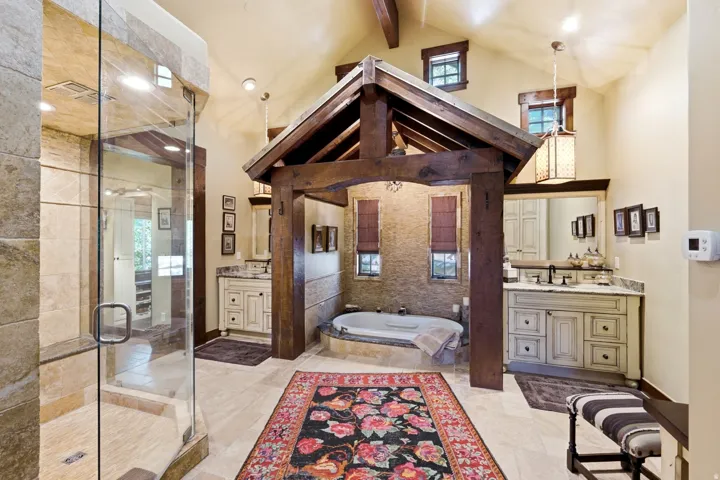 Expansive bathroom featuring a prominent timber-frame structure over the soaking tub, a spacious walk-in shower with stone-tiled walls, dual vanity cabinets with stone countertops, and vaulted ceilings with exposed wood beams