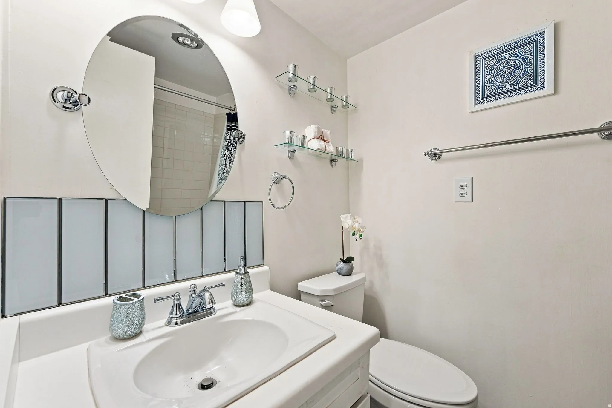 Vanity with integrated sink and chrome faucet, oval mirror, wall-mounted glass shelving, and a toilet