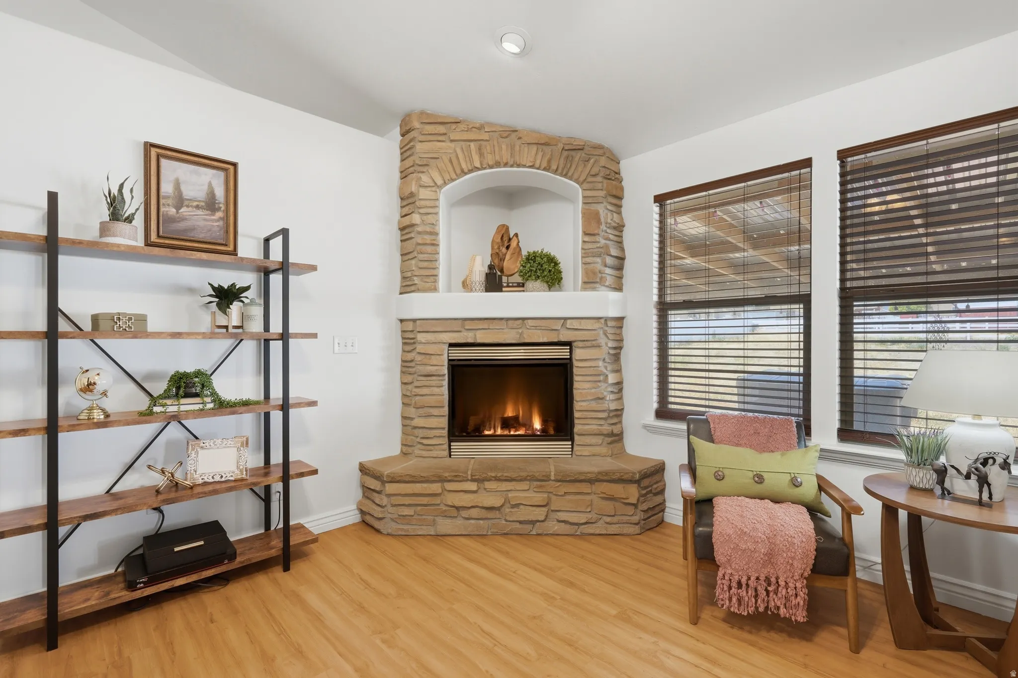 Corner fireplace with stone surround and hearth, wood-finish flooring, white painted walls, built-in shelving, and two windows with wood blinds