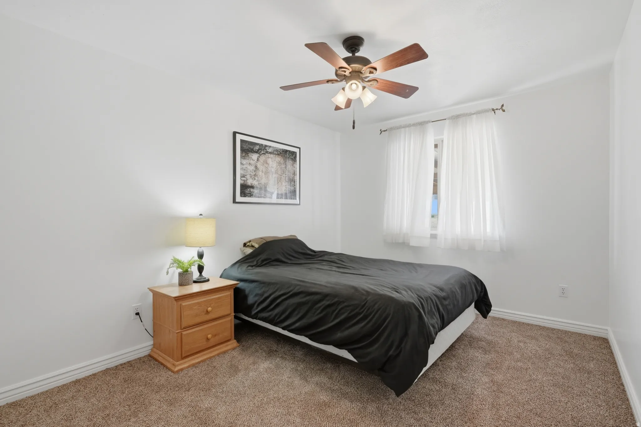 Carpeted bedroom featuring white walls, a window with sheer curtains, a ceiling fan with integrated lighting, and white baseboards