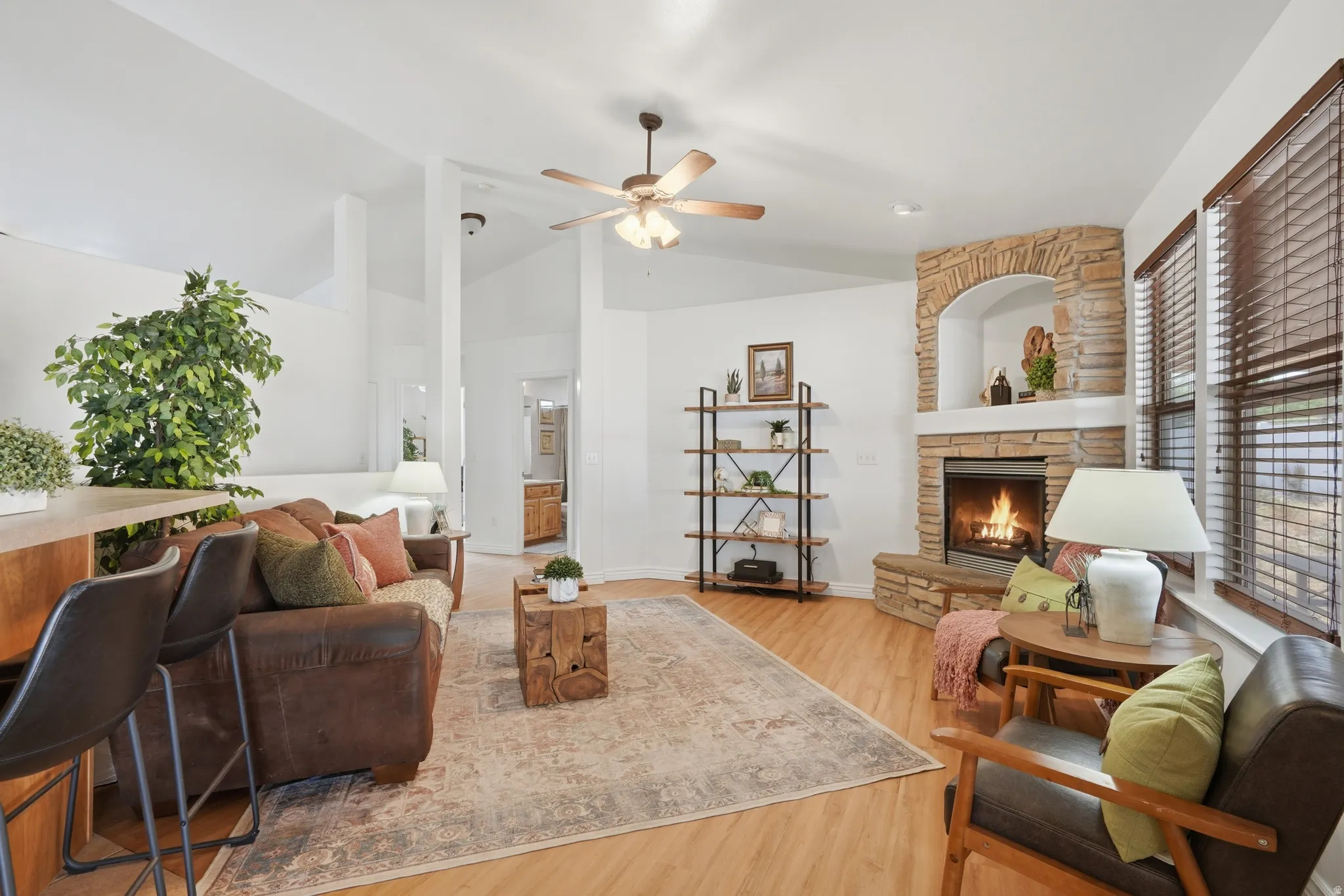 Spacious living area featuring vaulted ceilings, wood-finish flooring, a corner fireplace with stone surround, and multiple windows