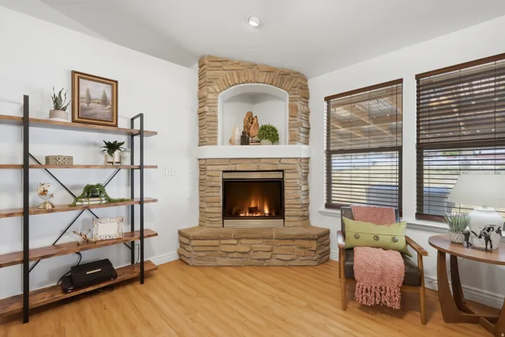 Corner fireplace with stone surround and hearth, wood-finish flooring, white painted walls, built-in shelving, and two windows with wood blinds