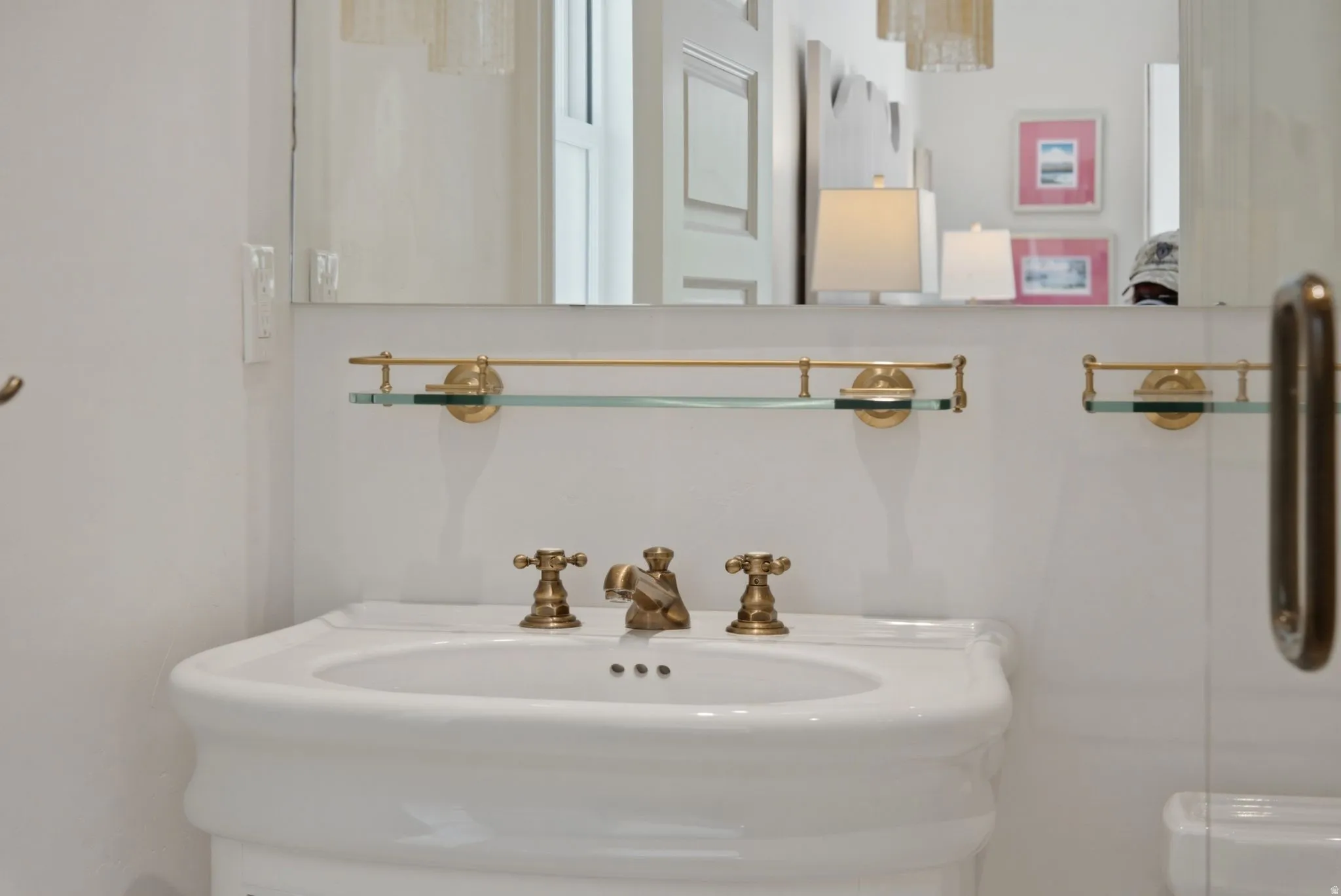 White pedestal sink with antique brass faucet and fixtures, featuring a wall-mounted glass shelf