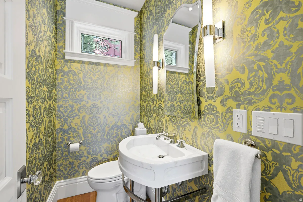Powder room featuring ornamental wallpaper, a corner pedestal sink with chrome faucet, a decorative mirror, and two vertical wall sconces