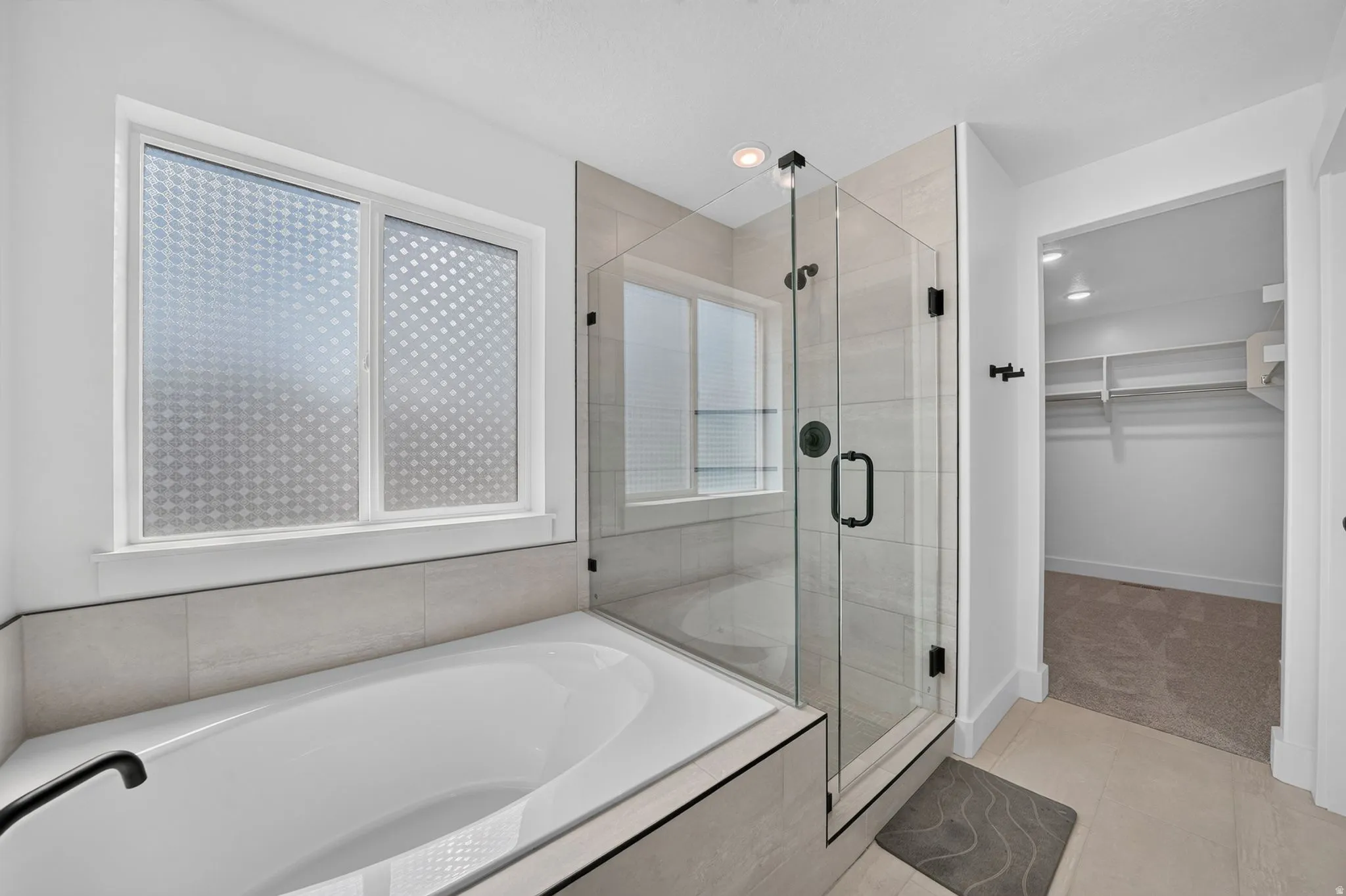 Contemporary primary bathroom featuring a built-in soaking tub with matte black fixtures, a glass-enclosed shower, and light-toned tile flooring