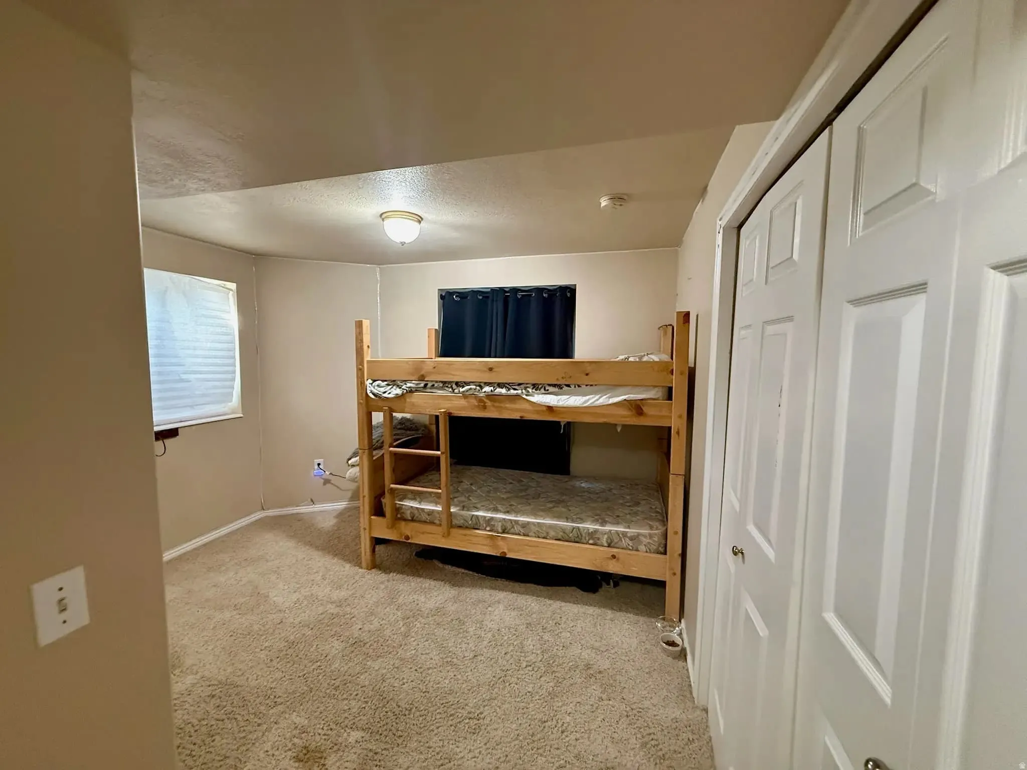 Carpeted room featuring a ceiling-mounted light fixture, white interior walls, a window with horizontal blinds, and a built-in closet with bifold doors