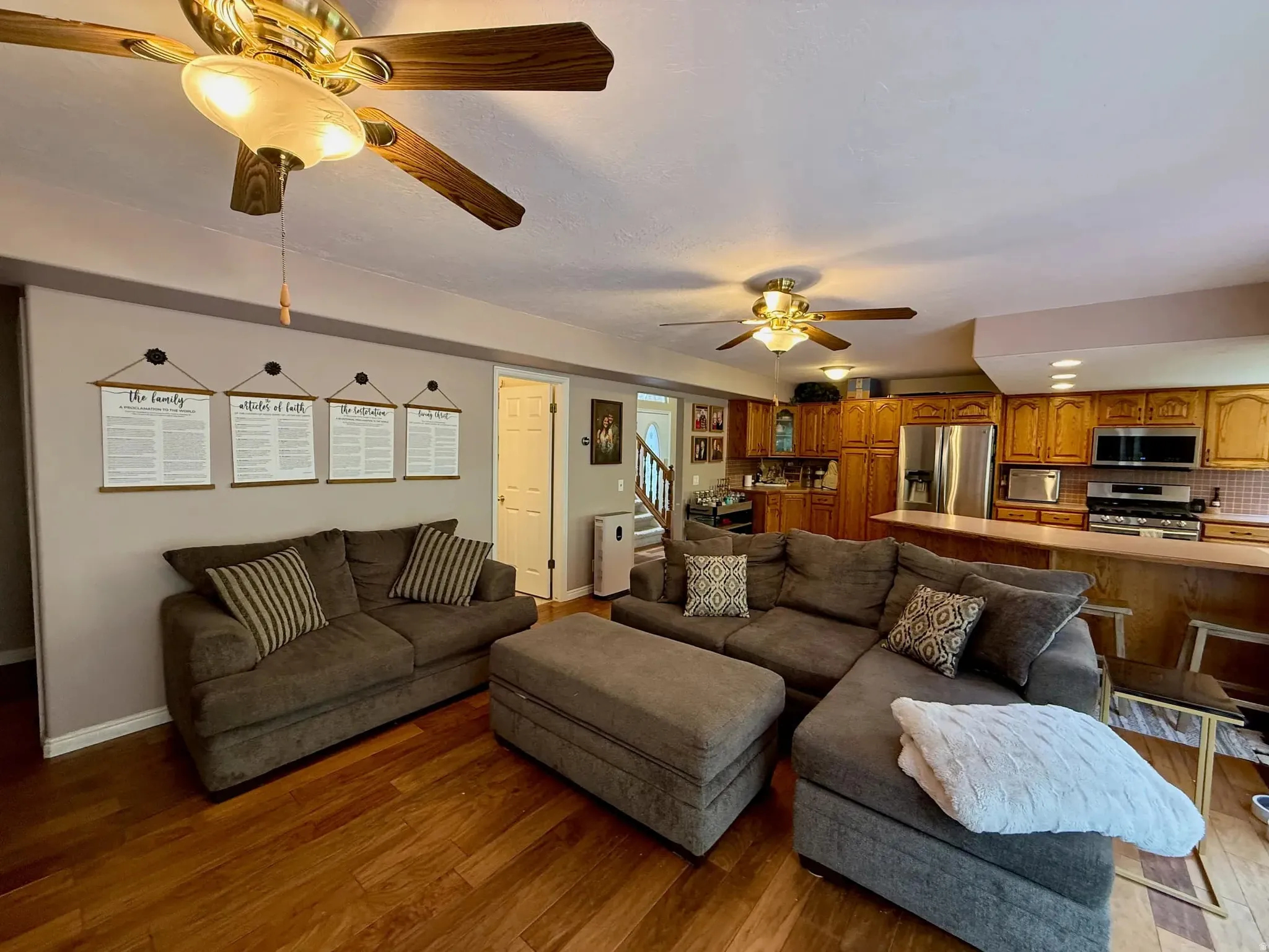 Open-concept living area featuring wood-finish flooring, two ceiling fans with integrated lighting, and a kitchen with warm wood cabinetry and stainless steel appliances