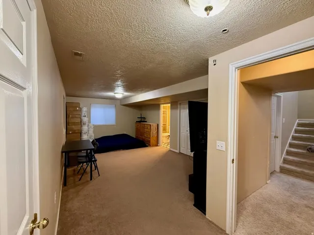 Expansive carpeted room featuring a recessed light fixture, a ceiling-mounted light fixture, a window with a blind, and a doorway leading to a staircase