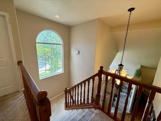 Carpeted staircase with wood-finish balusters and handrails, featuring an arched window with detailed muntins, a recessed ceiling light, and a multi-bulb hanging chandelier