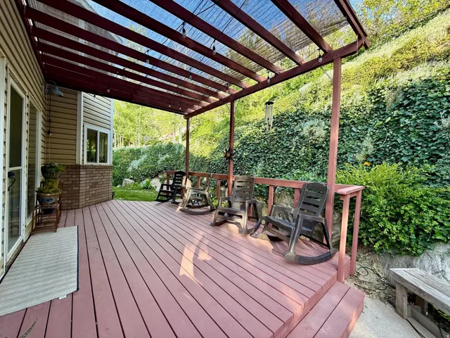 Expansive outdoor deck with a pergola and sunshade, featuring wood-finish flooring and a surrounding natural rock and foliage landscape