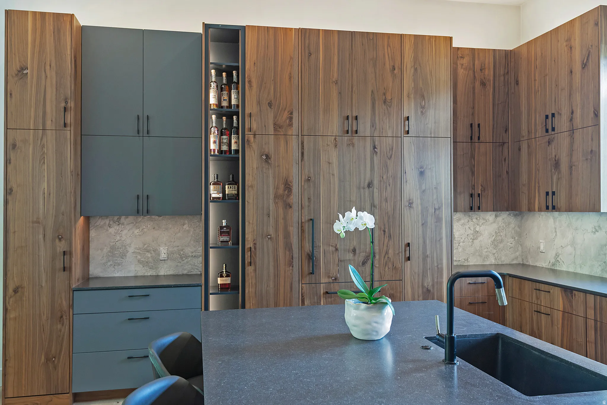 Contemporary kitchen featuring custom wood-finish cabinetry with integrated hardware, a dual-tone design with slate blue lower cabinets, and a central island with waterfall-edge countertop