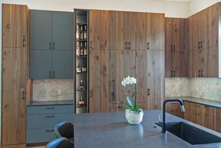Contemporary kitchen featuring custom wood-finish cabinetry with integrated hardware, a dual-tone design with slate blue lower cabinets, and a central island with waterfall-edge countertop
