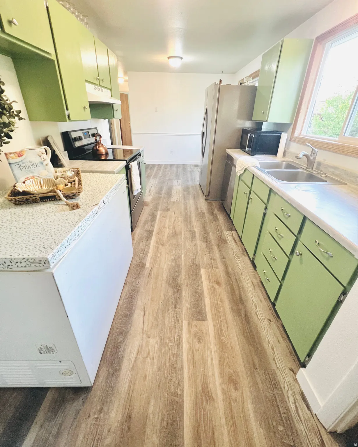 Galley kitchen featuring wood-finish flooring, green cabinetry, stainless steel appliances, a double basin sink, and a built-in oven with overhead microwave