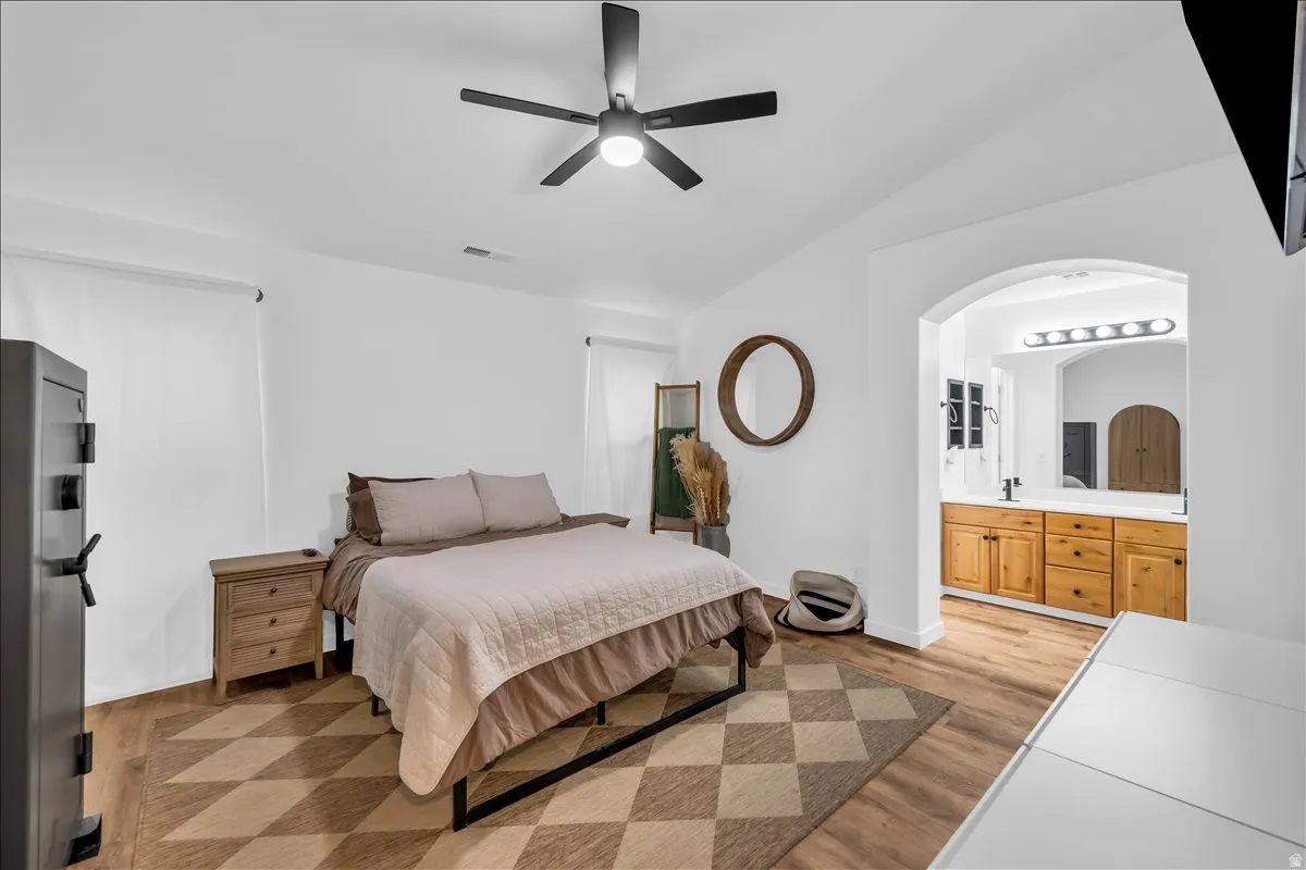 Bedroom featuring wood-finish flooring, a ceiling fan, and a wide arched doorway leading to a bathroom with a dual vanity, light countertop, and wood cabinetry