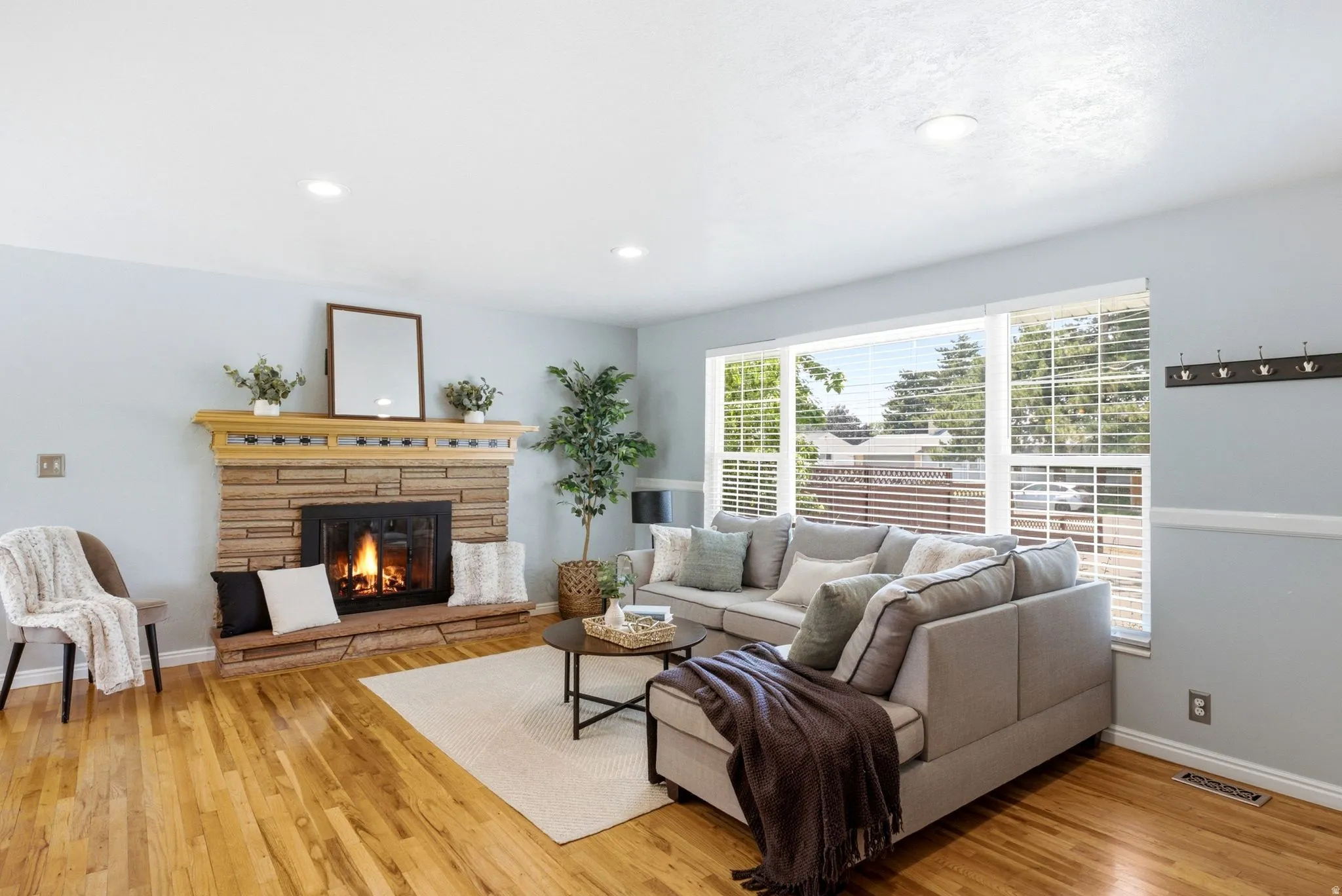 Living room with a stone-clad fireplace and hearth, wood-finish flooring, a large picture window with blinds, and recessed lighting