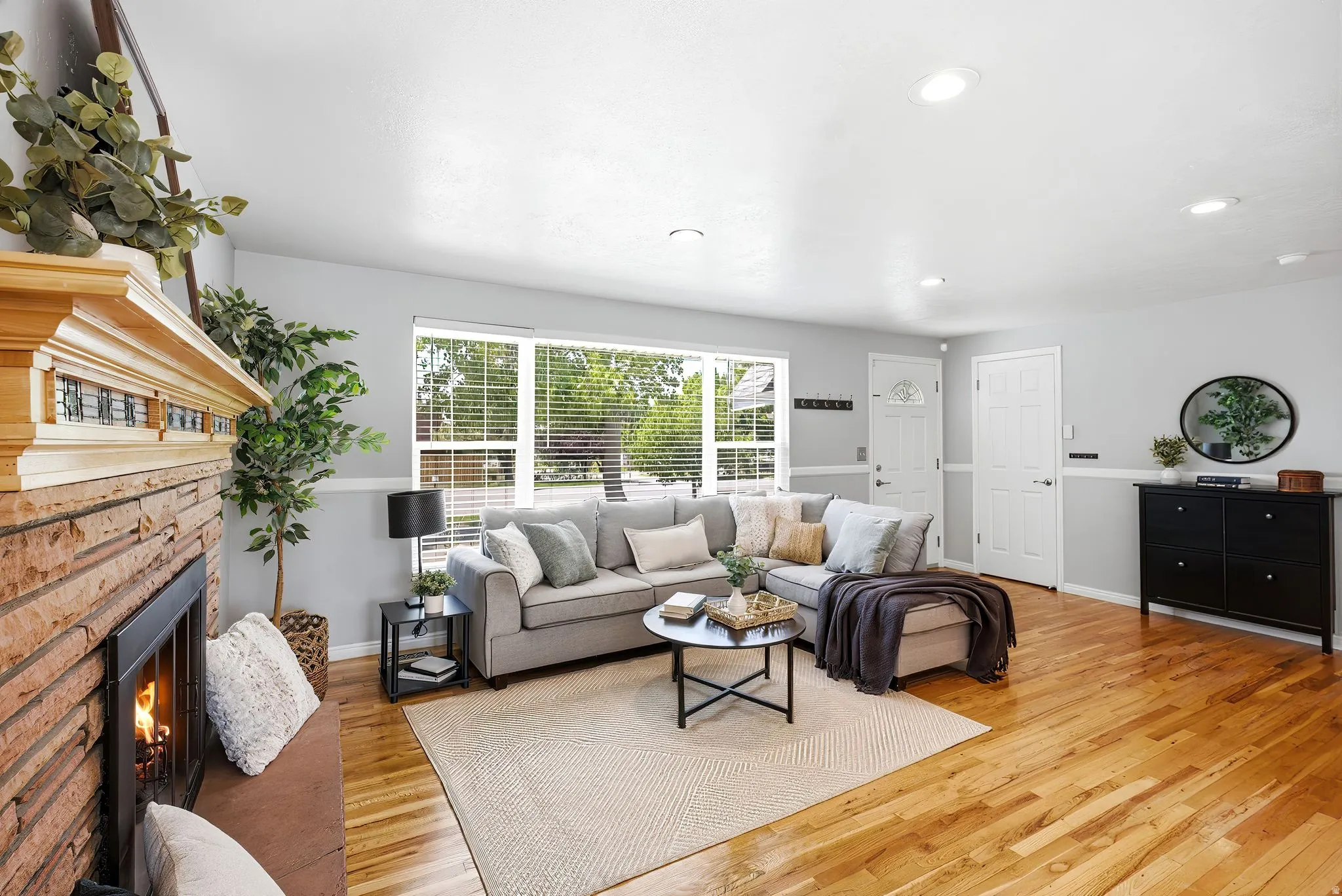 Spacious living area featuring wood-finish flooring, a brick fireplace with a built-in hearth, and abundant natural light through large windows