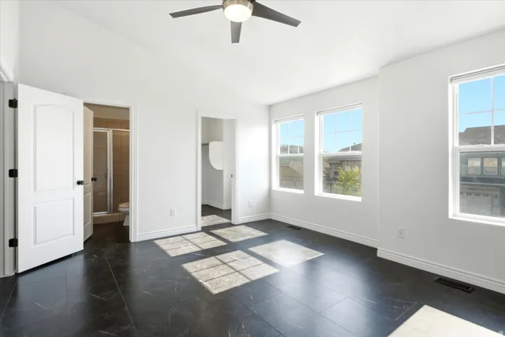 Bright interior featuring dark tile flooring, white walls, multiple windows, a ceiling fan, and access to an ensuite bathroom with a glass-enclosed shower