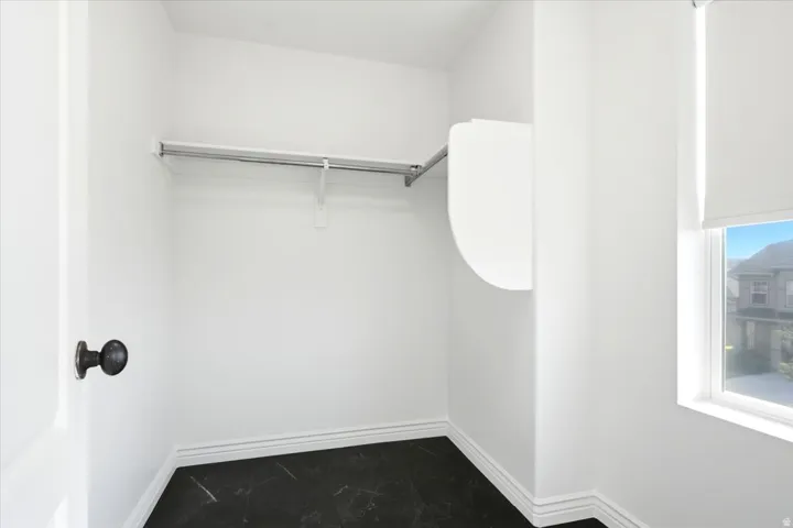 Walk-in closet featuring a single hanging rod, black marble-look flooring, white baseboards, and a window with a roller shade