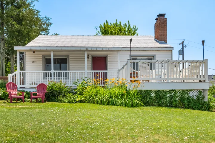 Exterior featuring horizontal siding, a red entry door, an elevated deck with white railing, a brick chimney, and a landscaped front yard with grass and plantings