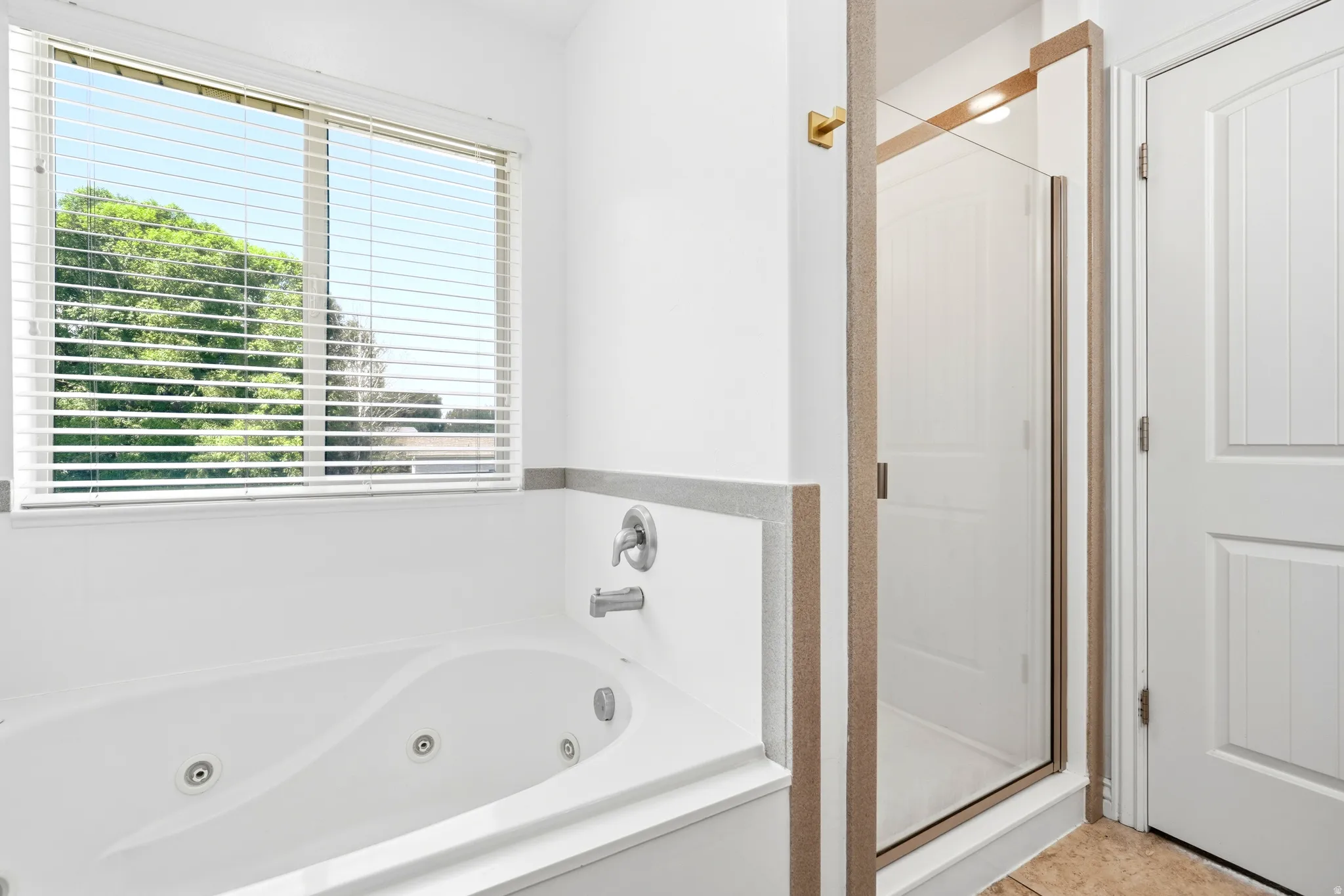 White jetted soaking tub with chrome fixtures, a large window featuring horizontal blinds, and a glass-enclosed shower with brushed gold trim