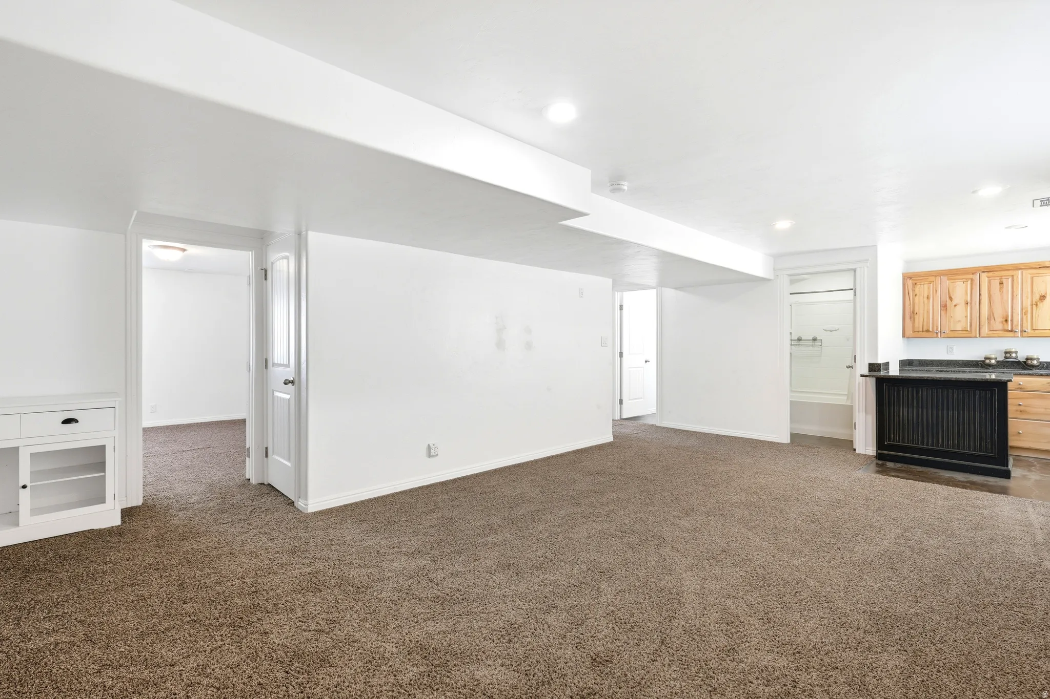 Spacious carpeted room featuring a white built-in cabinet with glass-front storage, recessed lighting, and a partial kitchen area with wood cabinetry and a dark countertop