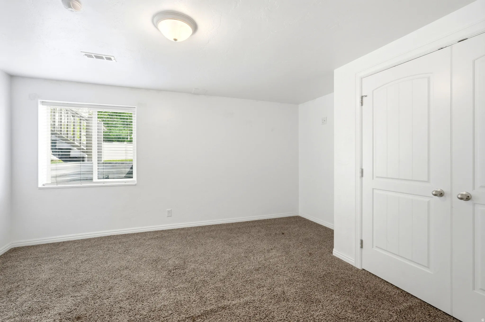 Carpeted room with white walls, a single window with blinds, and a double-door closet