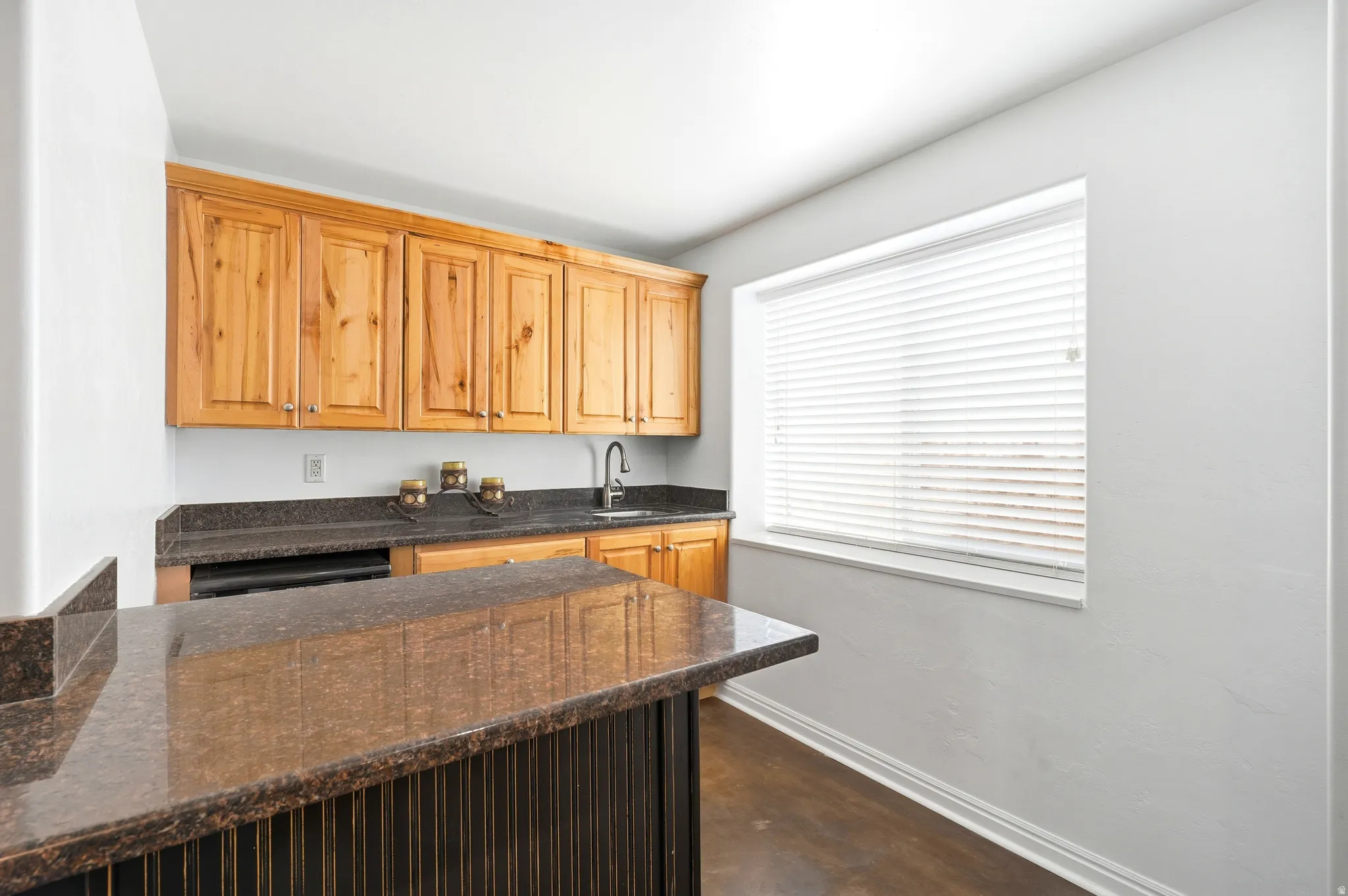 Kitchenette featuring wood cabinetry, dark speckled countertops, a built-in sink with a faucet, a window with blinds, and wood-finish flooring