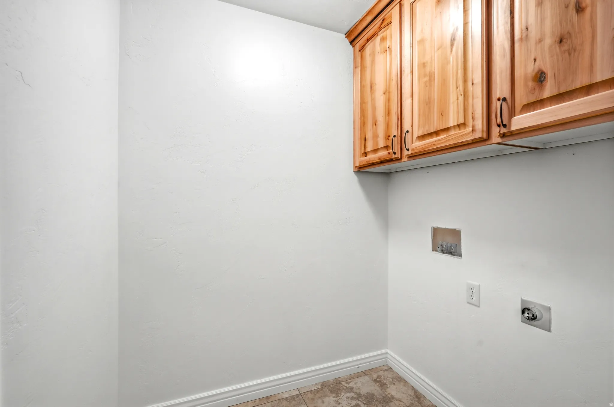 Functional laundry space featuring overhead wood cabinetry, tile flooring, baseboard trim, and utility hookups