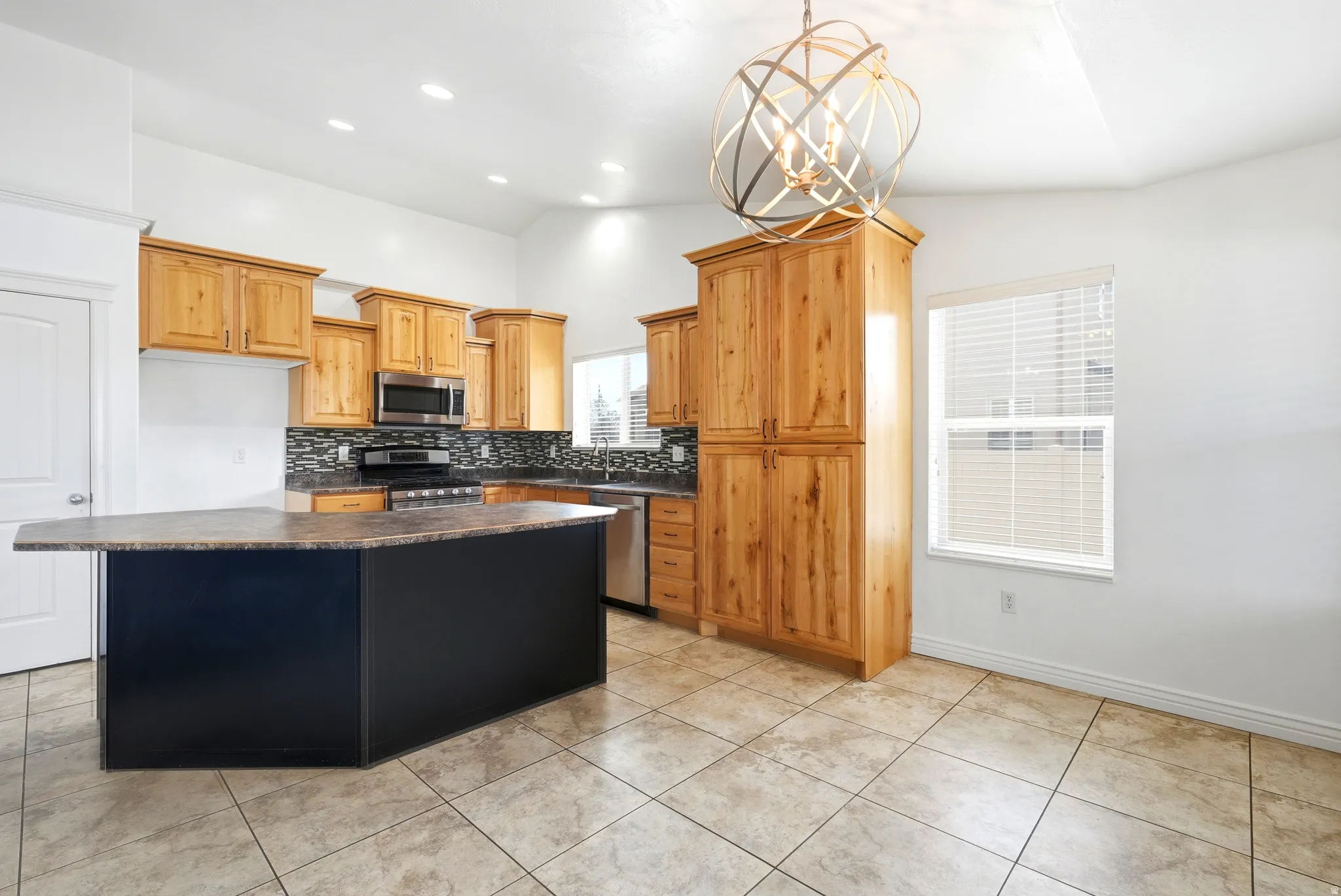 Kitchen featuring light wood cabinetry, a dark central island with a contrasting countertop, a patterned tile backsplash, stainless steel appliances, and tile flooring