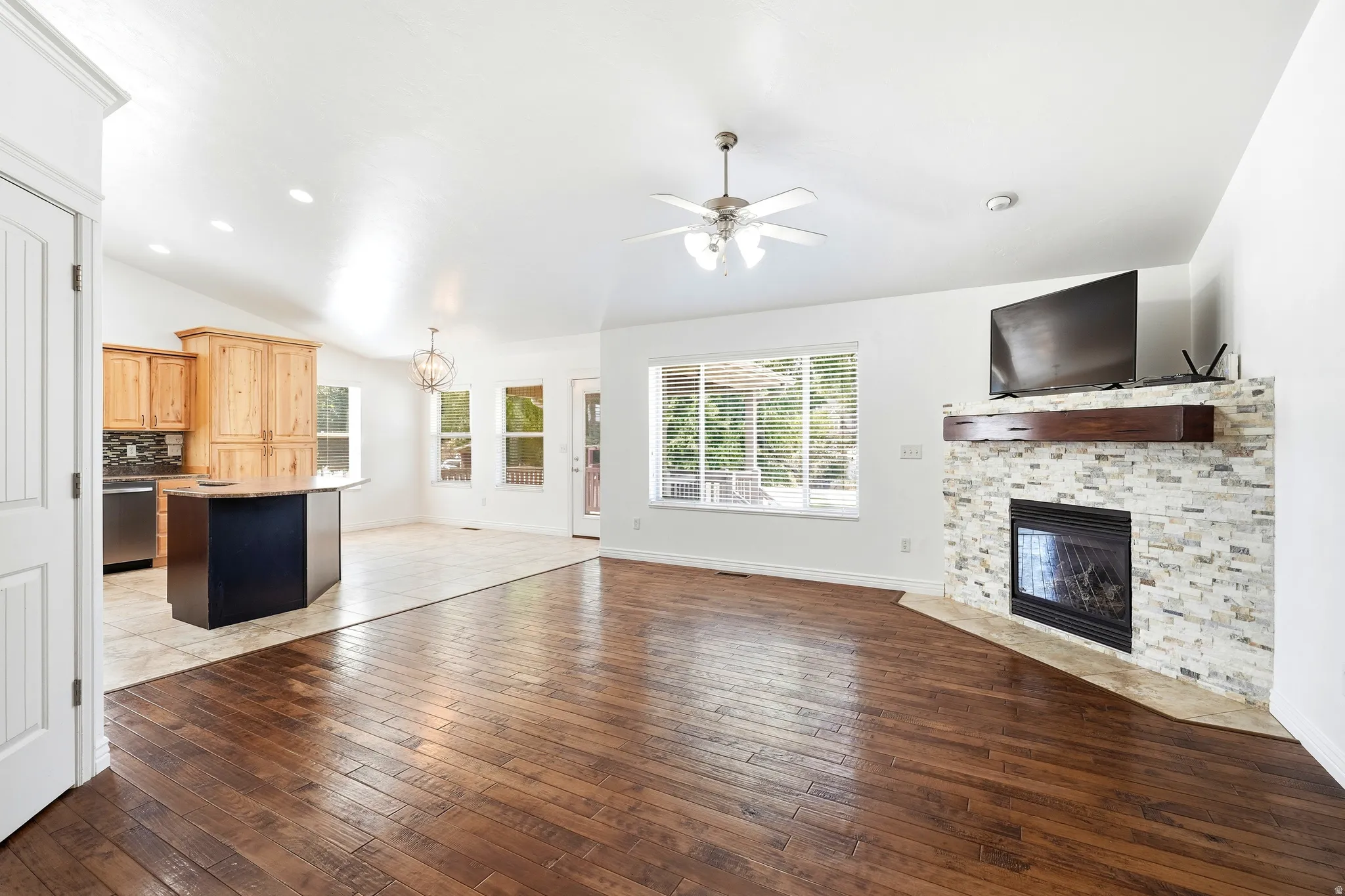 Great room featuring wood-finish flooring, a stone-surround fireplace with a wood mantle, and an open-concept kitchen with light wood cabinetry and a dark island