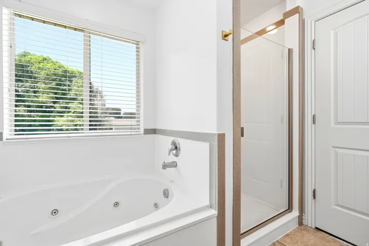 White jetted soaking tub with chrome fixtures, a large window featuring horizontal blinds, and a glass-enclosed shower with brushed gold trim