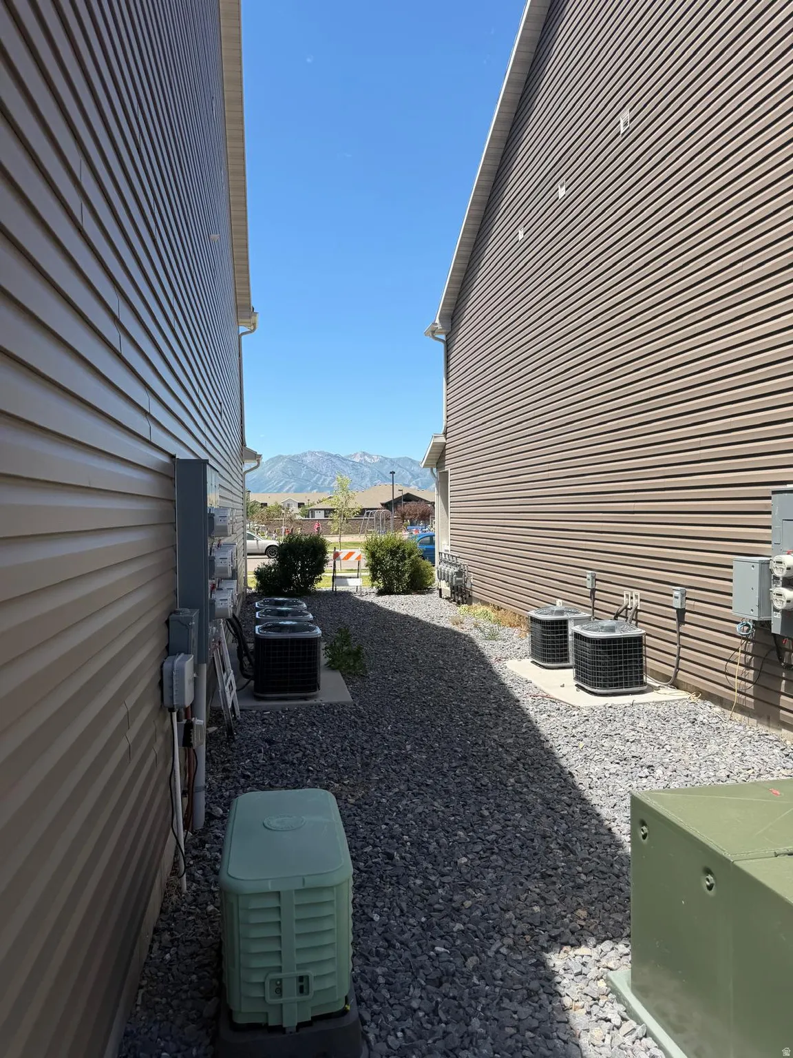 Exterior brown siding with light soffits and gutter system, featuring a gravel side yard with multiple HVAC units and electrical meters