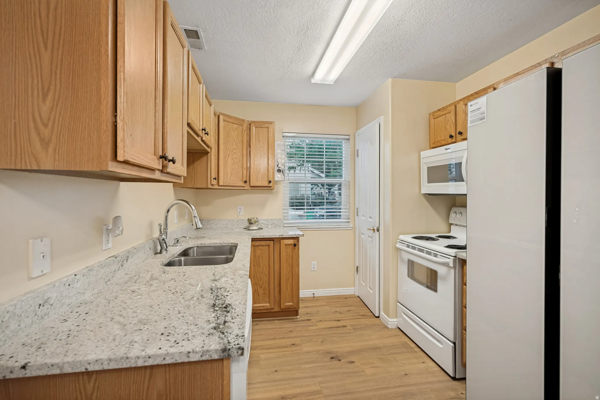 Kitchen featuring light wood cabinetry, light-toned granite-style countertops, double basin stainless steel sink with gooseneck faucet, white appliances, and wood-finish flooring