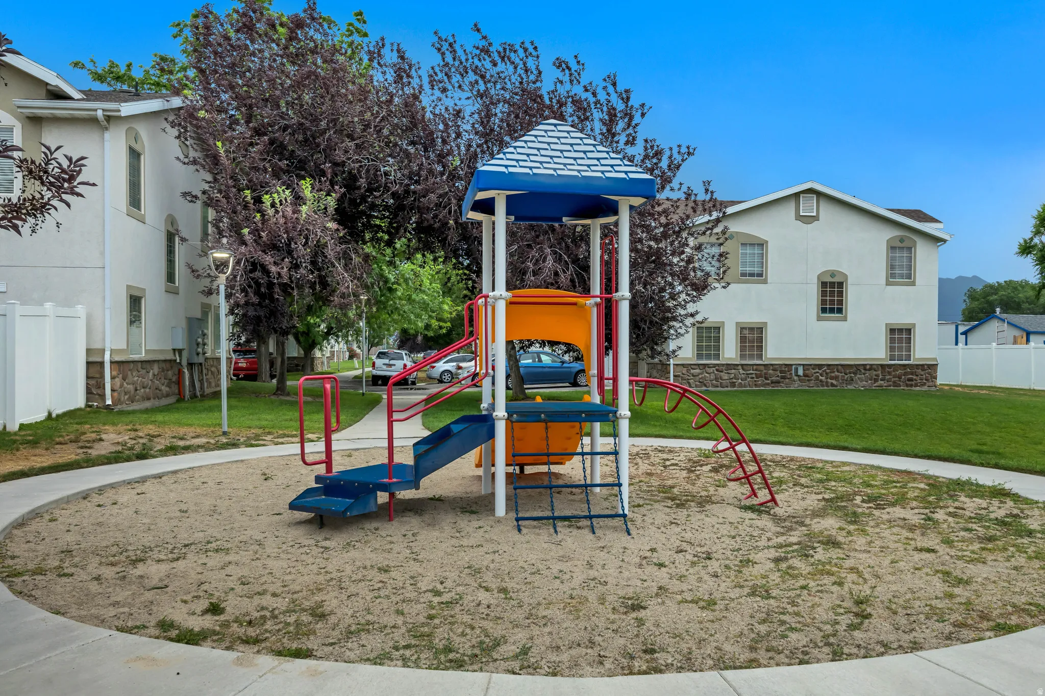 Recreational area featuring a multi-colored play structure, sandy ground cover, and surrounding concrete path
