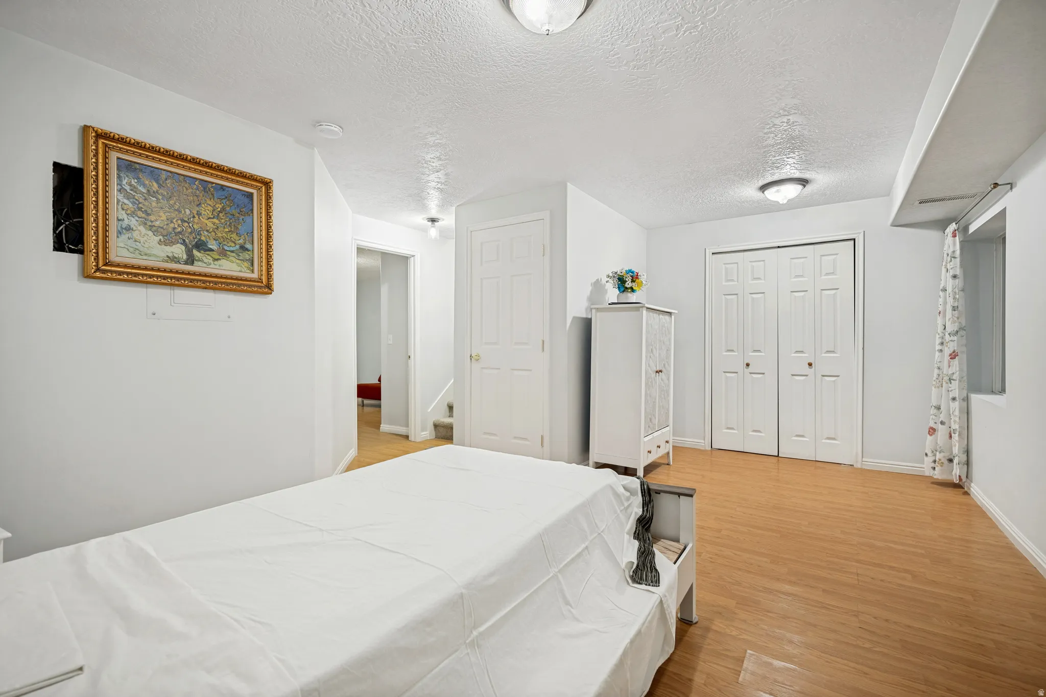 Light wood-finish flooring throughout, featuring a white paneled door, a bi-fold closet with paneled doors, and a window with a curtain rod