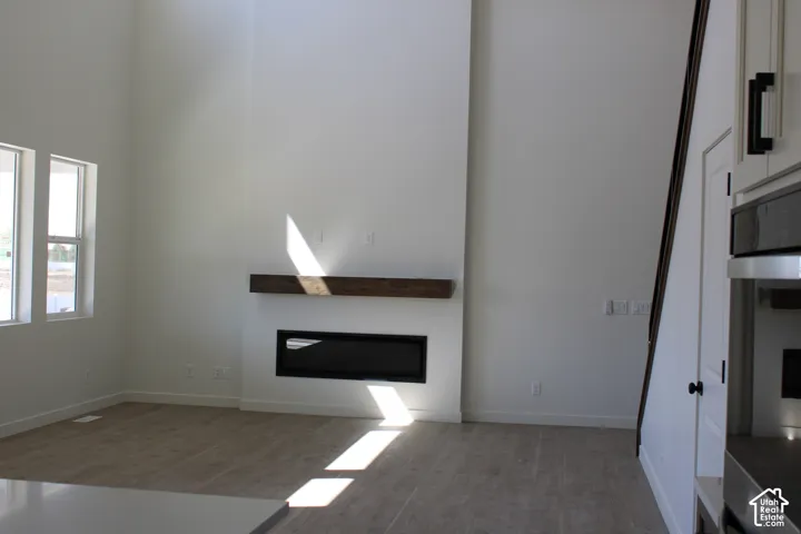 Double-height living area featuring a linear electric fireplace with a substantial wood mantel, two large windows, wood-finish flooring, and white wall finishes