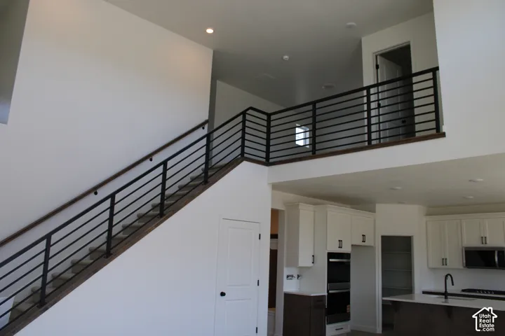 Open-concept interior featuring a two-story ceiling, modern horizontal metal stair railing with wood handrail, recessed lighting, and a kitchen with white cabinetry, built-in double ovens, and a dark island base