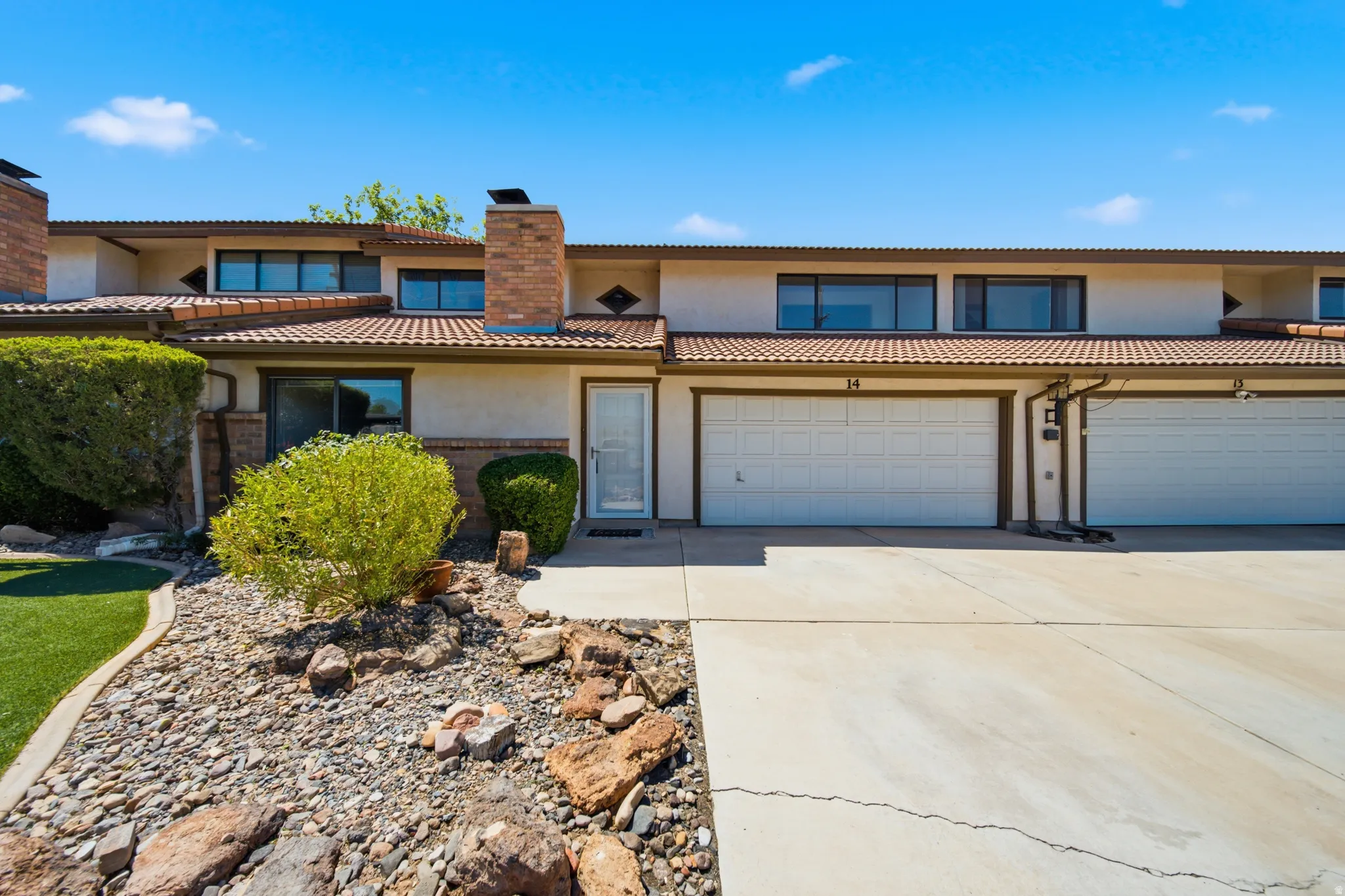 Stucco exterior with a terracotta-style tile roof, integrated two-car garage, brick chimney, and desert-style landscaping