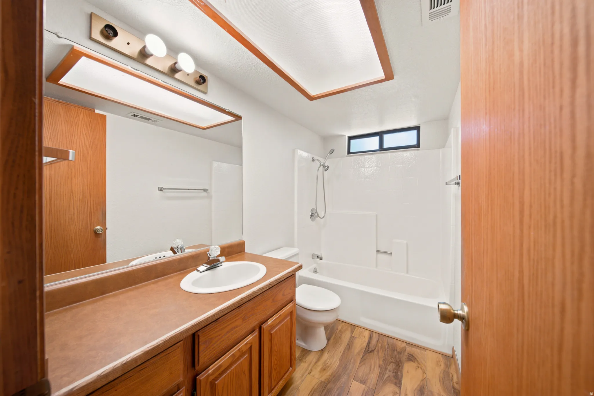 Bathroom featuring a wood-finish vanity with a built-in sink, a large mirror, a shower-tub combination with a white surround, and wood-finish flooring