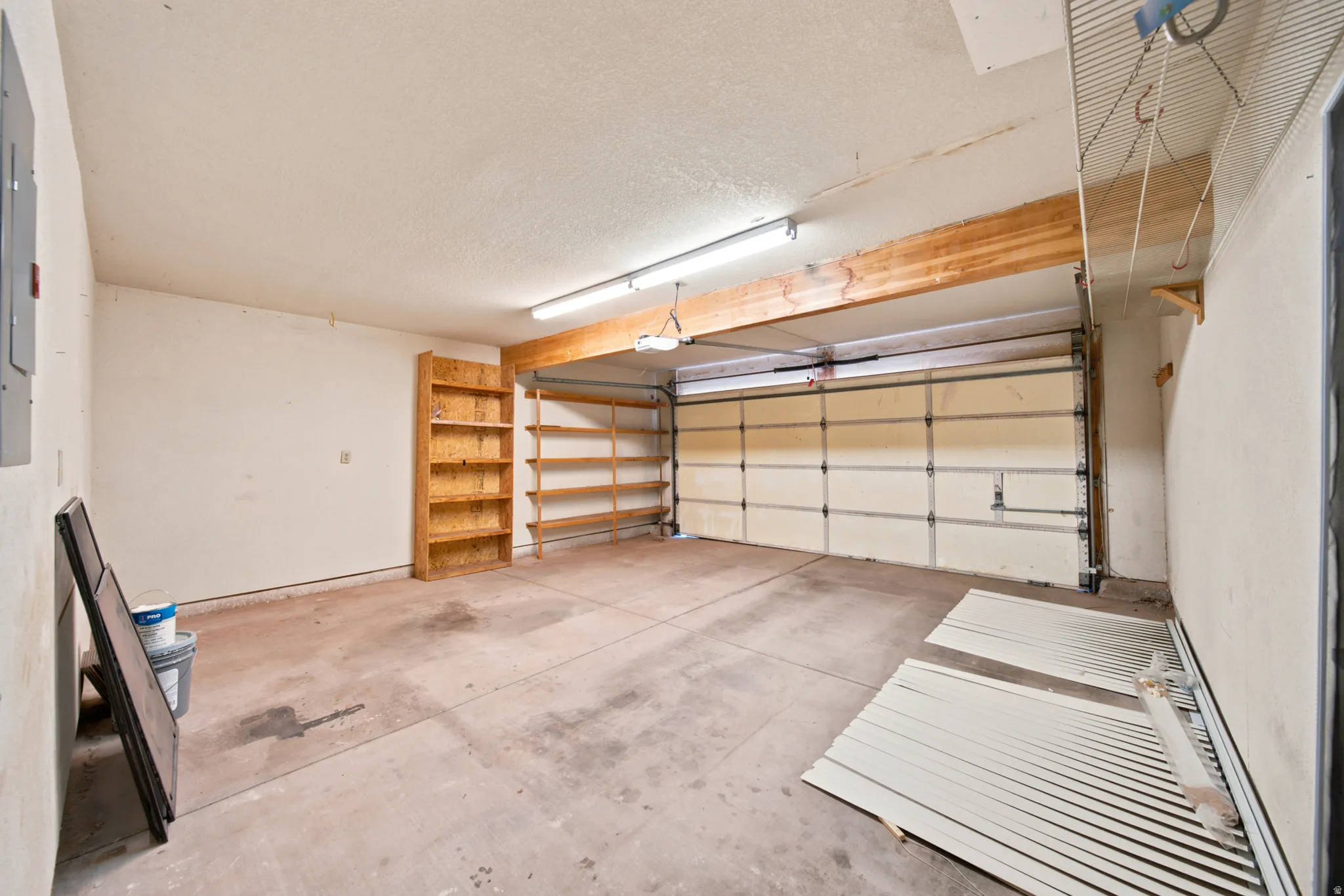 Single-bay garage with overhead door, concrete flooring, built-in wooden shelving, exposed timber beam, and fluorescent lighting