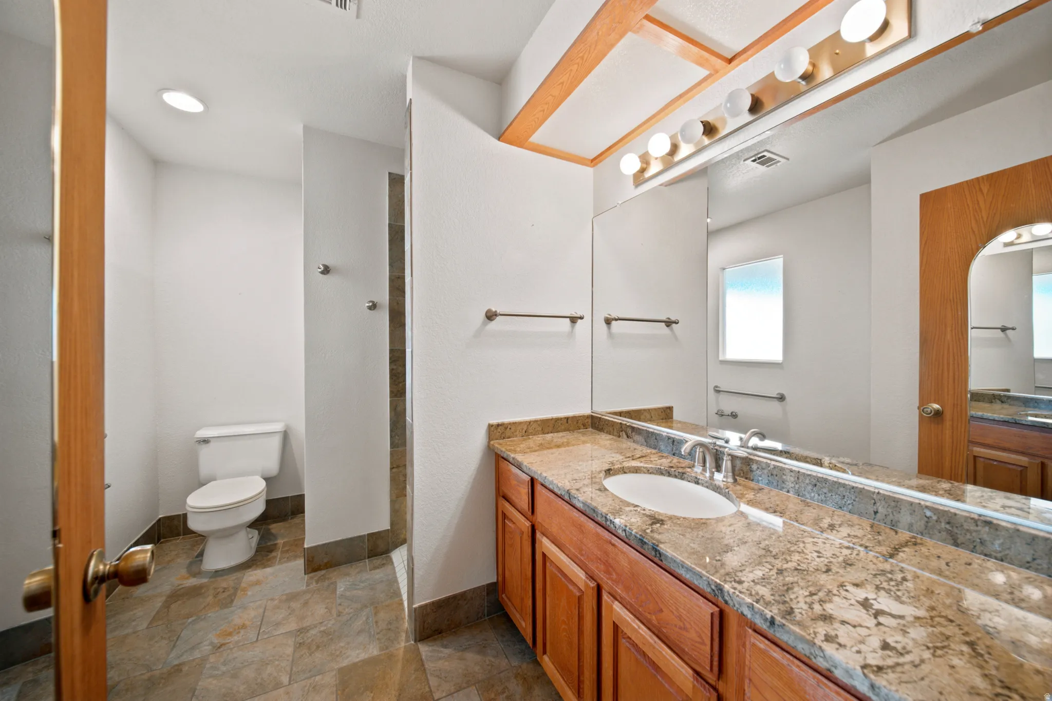 Bathroom featuring a stone-look tiled floor, a vanity with wood cabinetry, a light-toned granite countertop, a single undermount sink, and an expansive wall mirror