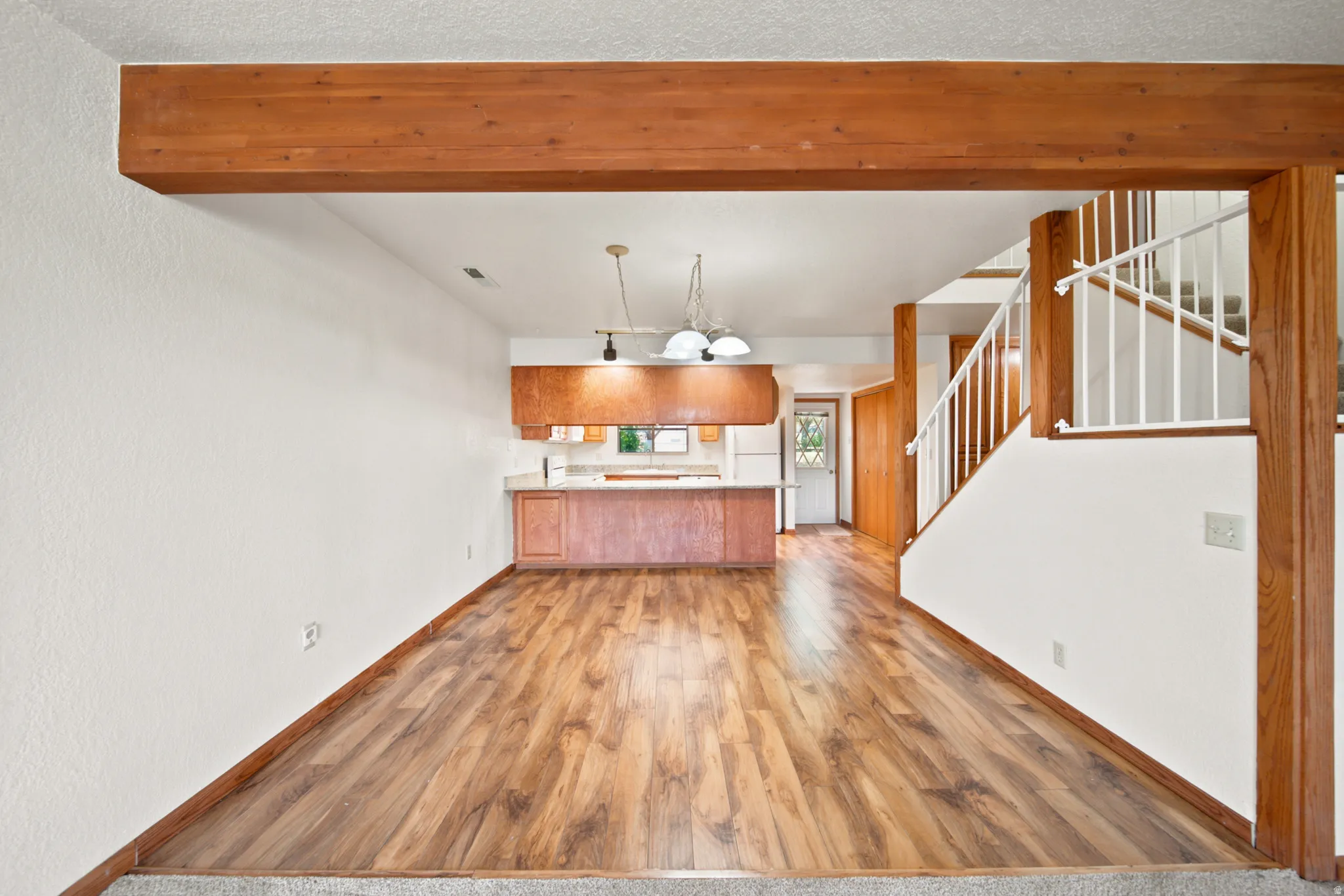 Open-concept living area featuring wood-finish flooring, a prominent exposed wood beam, a visible staircase with white balusters, and a kitchen area with wood cabinetry and light countertops