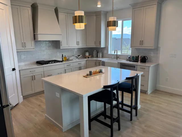 Modern kitchen featuring light wood-finish flooring, a large central island with seating, light gray cabinetry, a white farmhouse-style sink, and contemporary pendant lighting fixtures